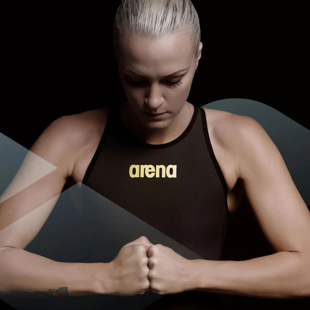 Woman in a black Arena high-neck swimsuit holds her hands together against a black background.