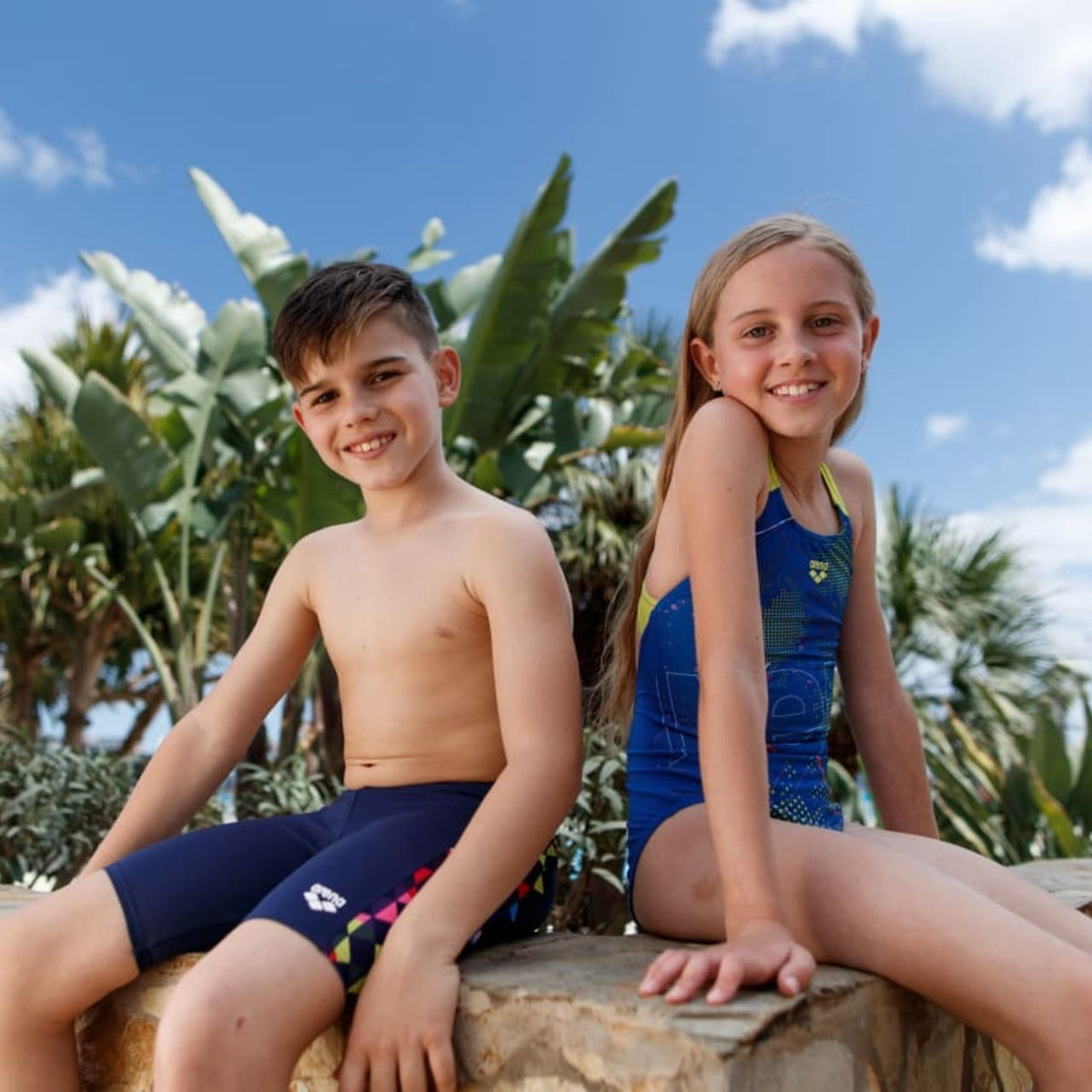 Boy in navy swim briefs and girl in a blue and green swimsuit sit together outdoors.