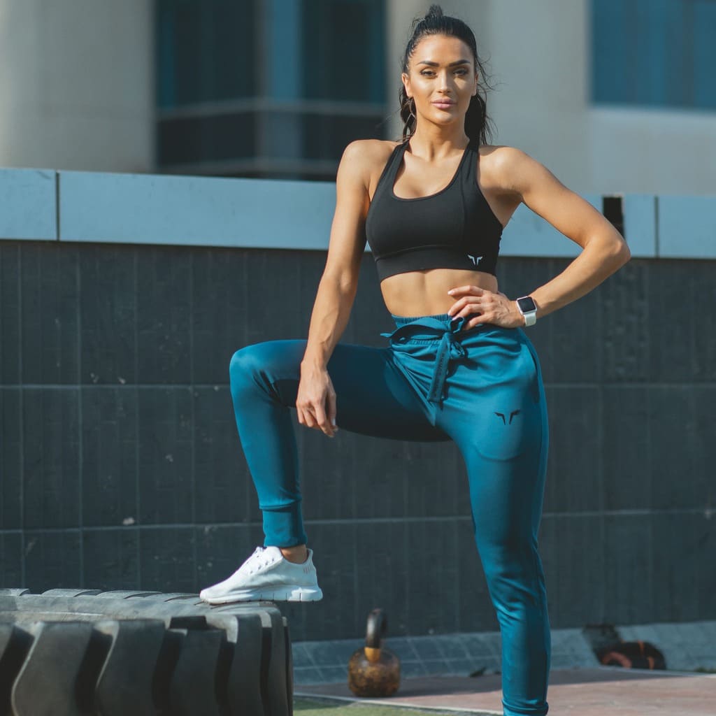 Woman wearing teal Squat Wolf joggers and a black sports bra standing with one foot on a large tyre.