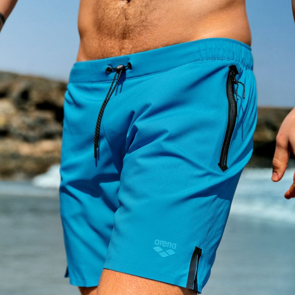 Close-up of a man wearing blue Arena swim shorts on a beach.