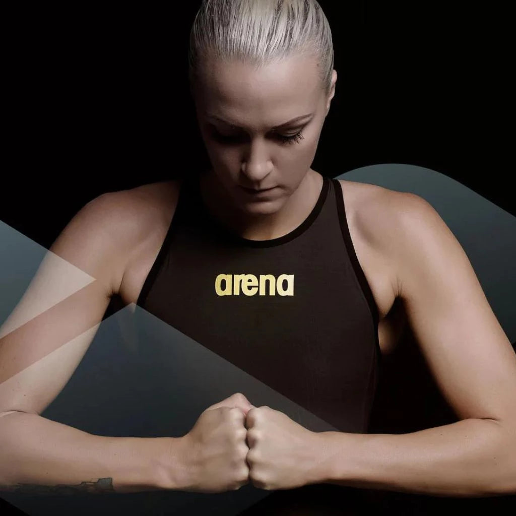 Woman wearing a black Arena swimsuit with a high neckline against a dark background.