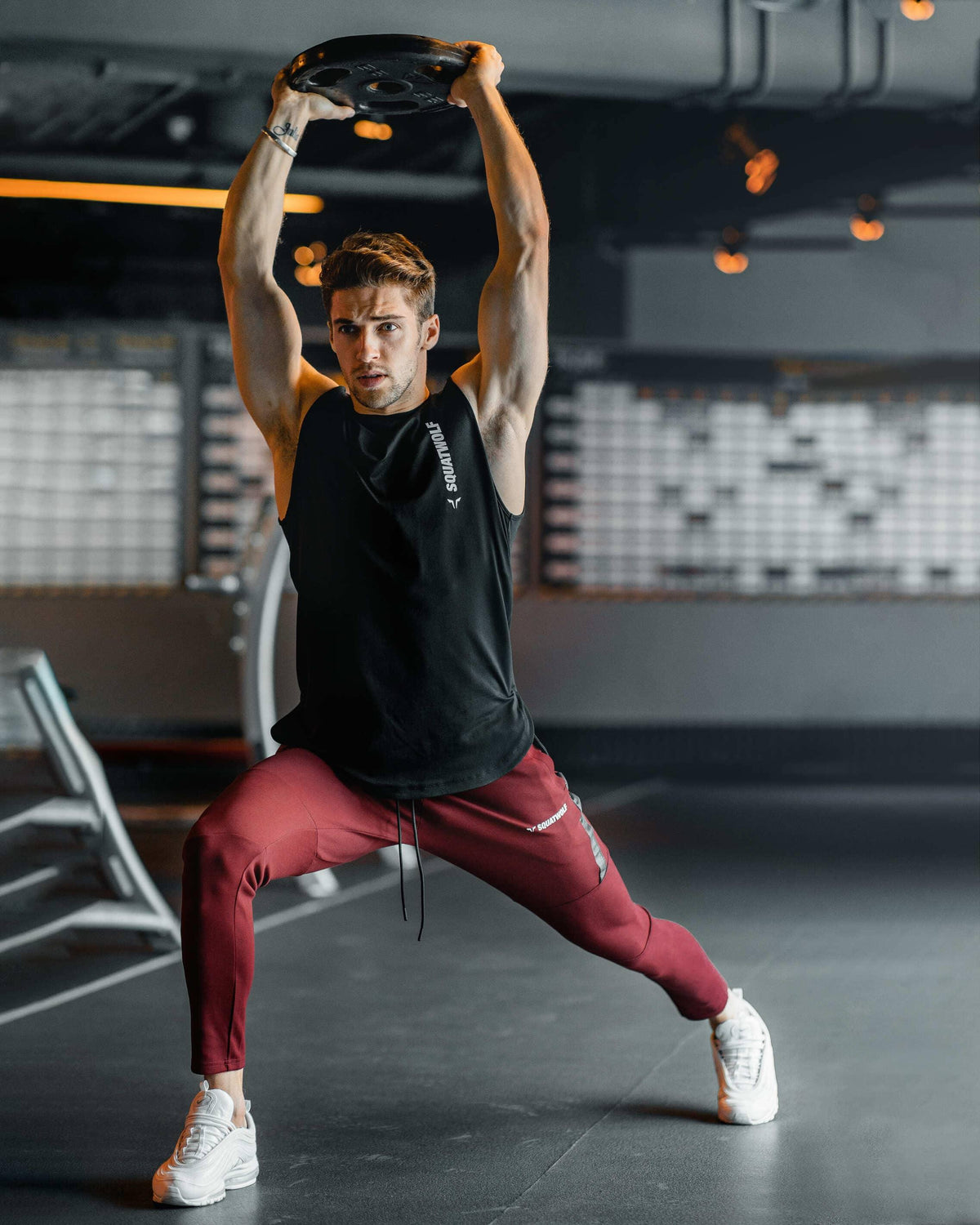Man wearing burgundy Squat Wolf joggers lunging while holding a weight plate overhead in a gym.