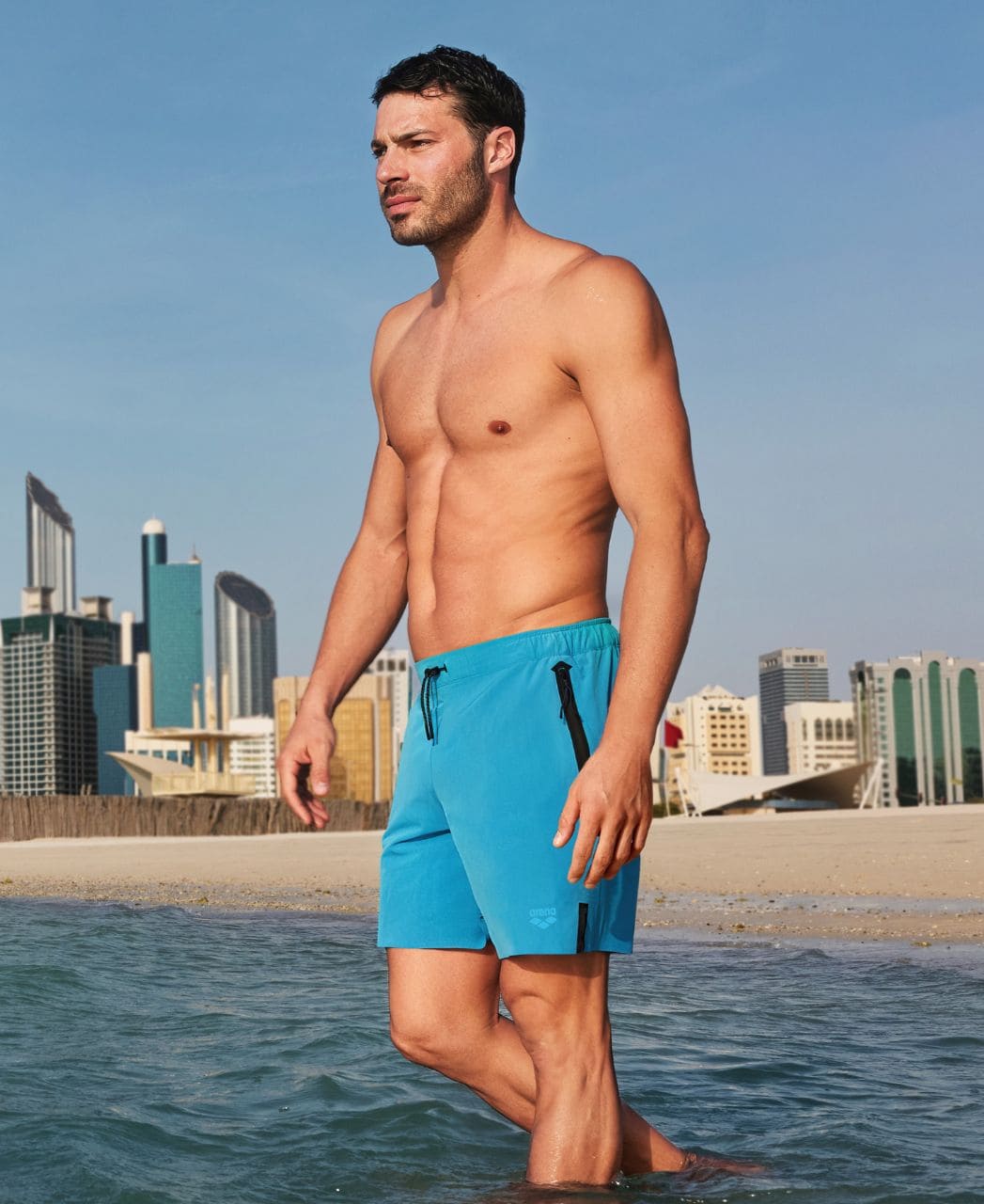 Herra stendur í sjónum í túrkísbláum Arena Evo Side Zip Pockets Beach Boxer sundbuxum með borg í bakgrunni