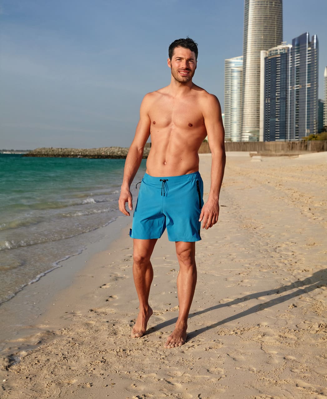 Herra gengur á strönd í túrkísbláum Arena Evo Side Zip Pockets Beach Boxer sundbuxum með borg í bakgrunni.