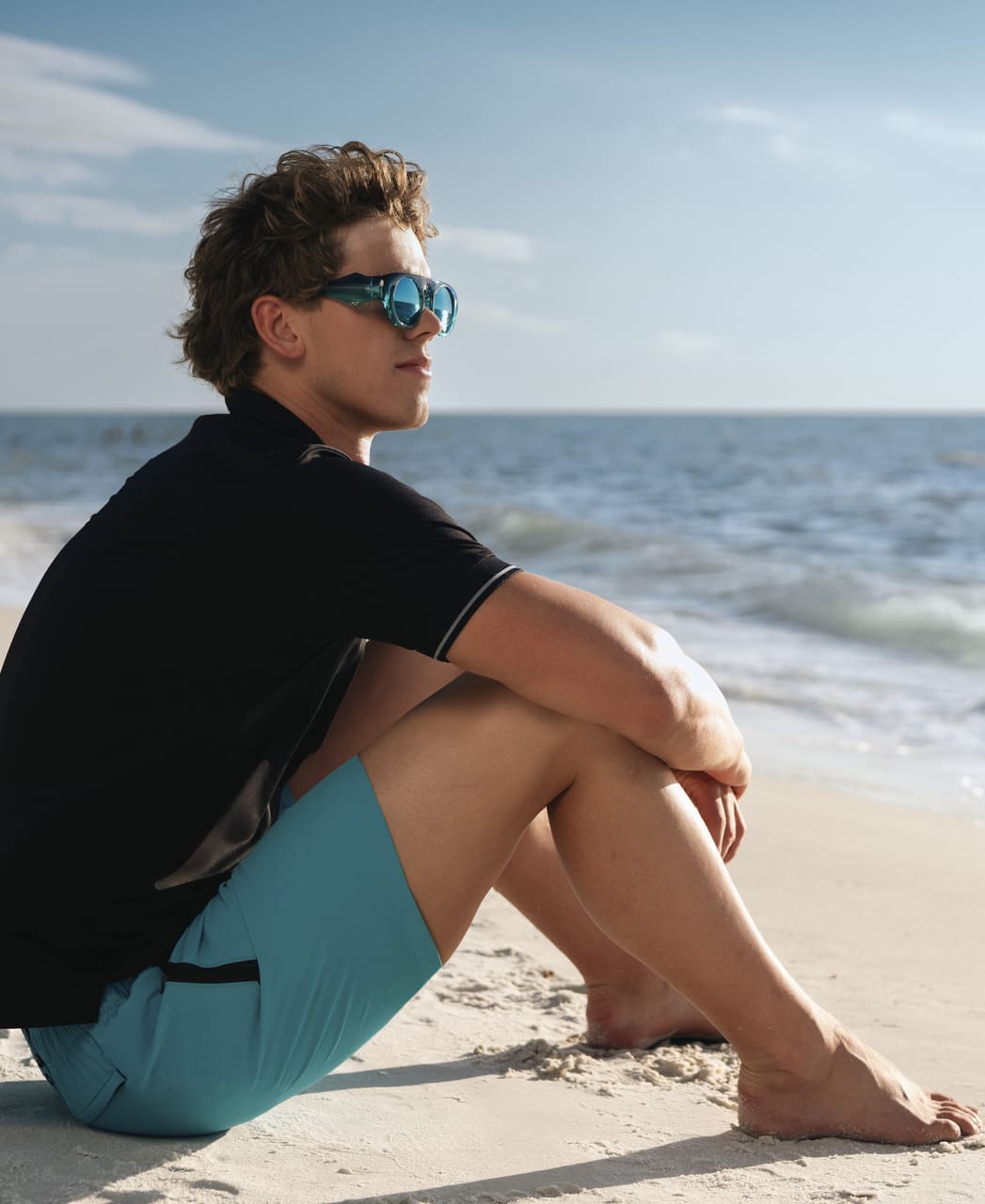 Karlmaður situr á strönd í túrkísbláum Arena Evo Side Zip Pockets Beach Boxer sundbuxum.