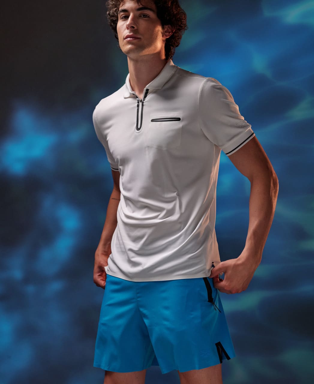 Karlmaður í hvítum Arena pólóbol og túrkísbláum Arena Evo Side Zip Pockets Beach Boxer sundbuxum.