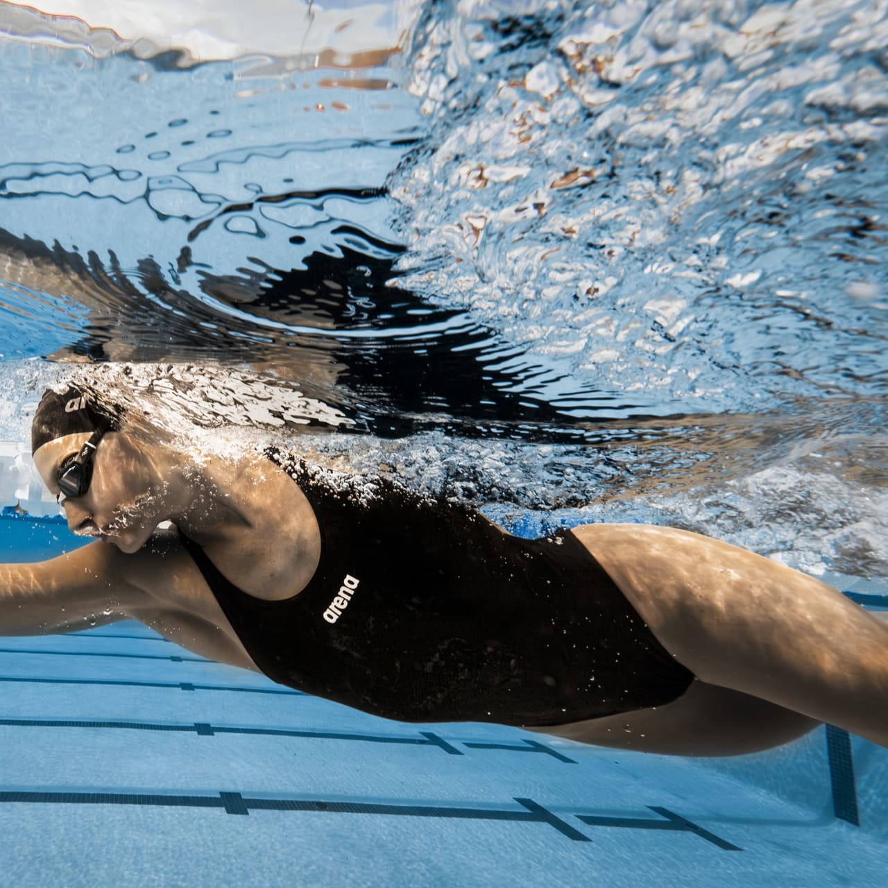 Myndin sýnir svartan Team sundbolinn fyrir konur úr Arena Performance línunni í notkun í sundlaug. Sundbolurinn er með góðu klórþoli úr MaxLife Eco sundfataefni Arena sem er framleitt úr 100% pólýester, að hluta til endurunnu pólýester. Lífstílsmynd sem sýnir konu á sundi í sundbolnum þar sem framhlið bolsins sést vel
