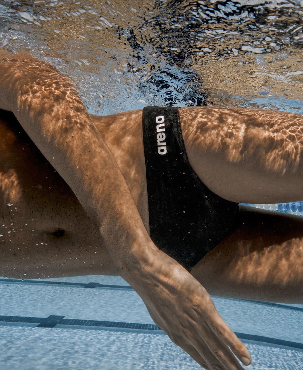 Nærmynd undir vatni af svartri Arena Team Swim Brief Solid sundskýlu með hvítu Arena lógói, MaxLife Eco efni sem heldur lögun vel við reglulega sundiðkun